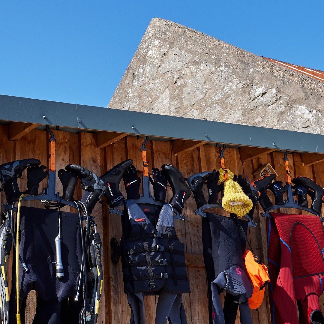 Wetsuits, gloves, and other diving gear hanging on specialized C-MONSTA hangers attached to a wooden outdoor wall | C-MONSTA